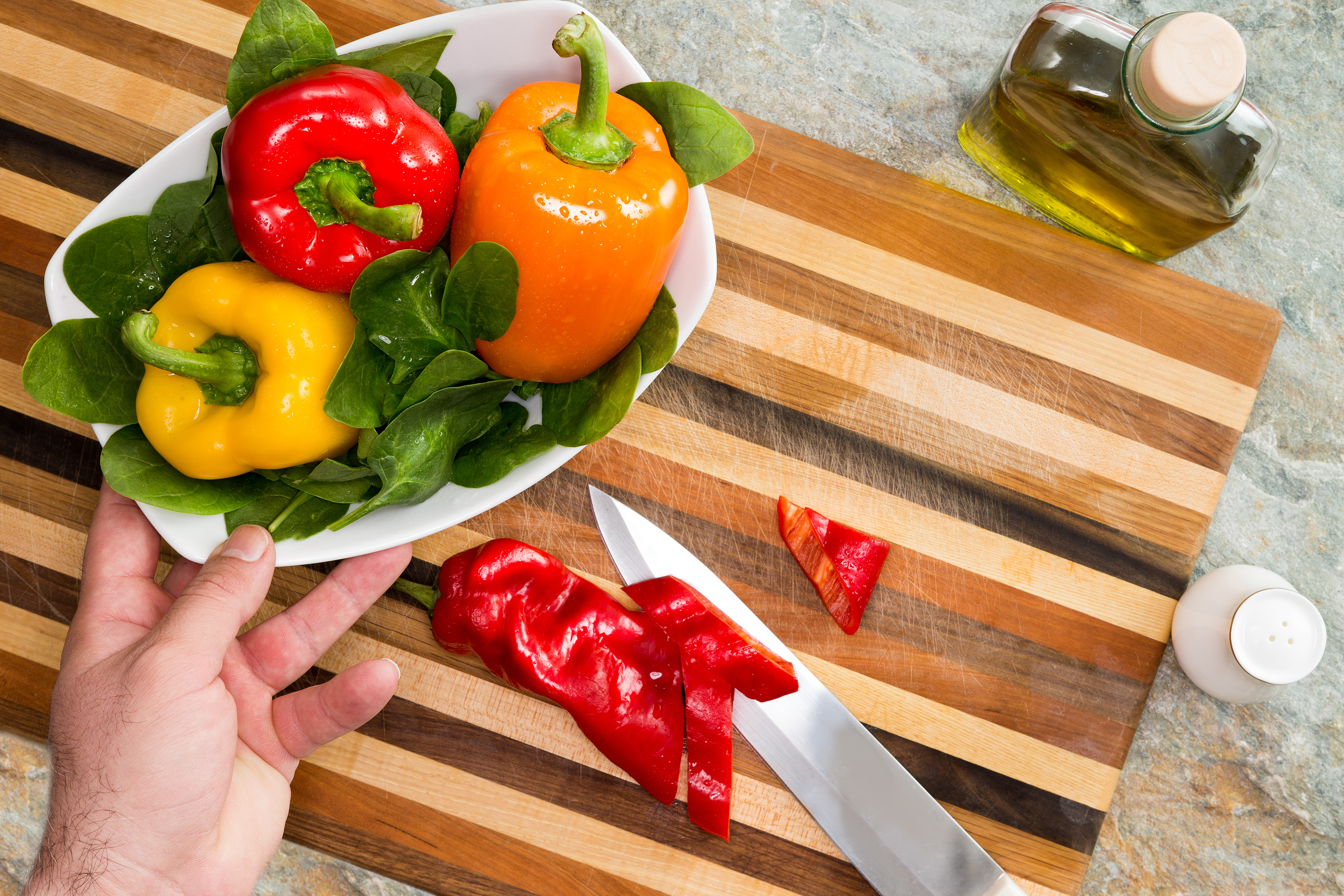 Man preparing a healthy fresh salad reaching for a bowl of colourful sweet bell peppers in red, yellow and orange with green baby spinach leaves as he chops the ingredients on a striped wooden board