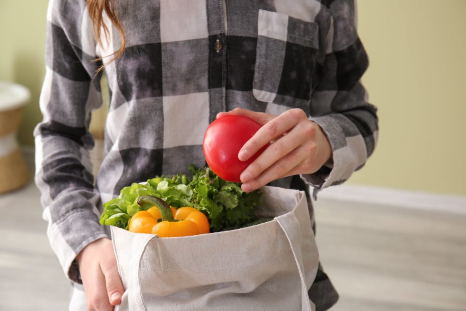 Woman placing Tomato into canvas shopping bag with other produce.